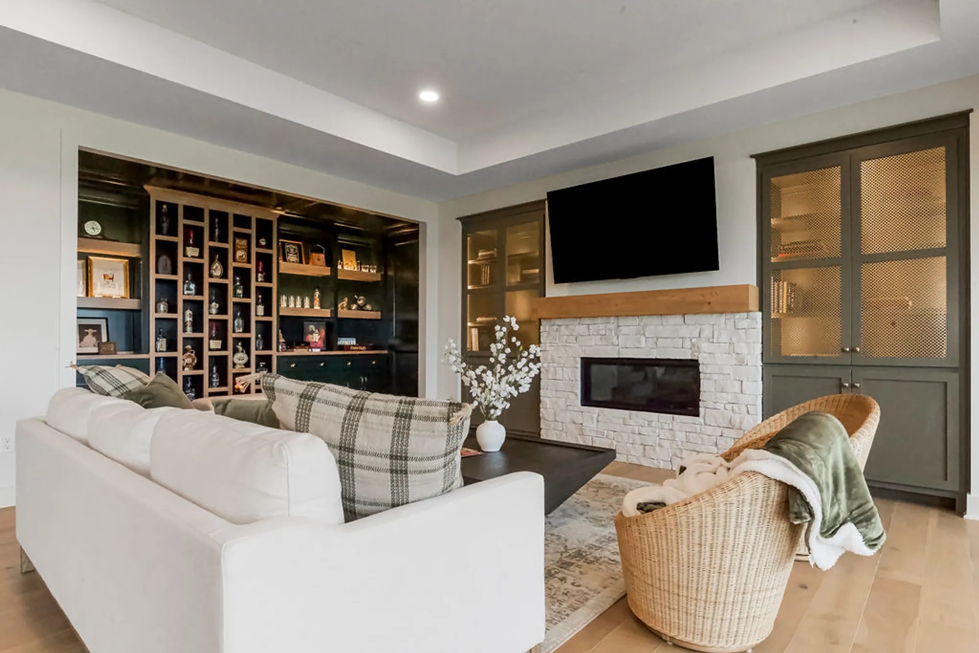 Bright open living room with a white sofa and plaid throw facing a stone fireplace, mounted TV above, and built-in shelves on the left filled with decor and books.