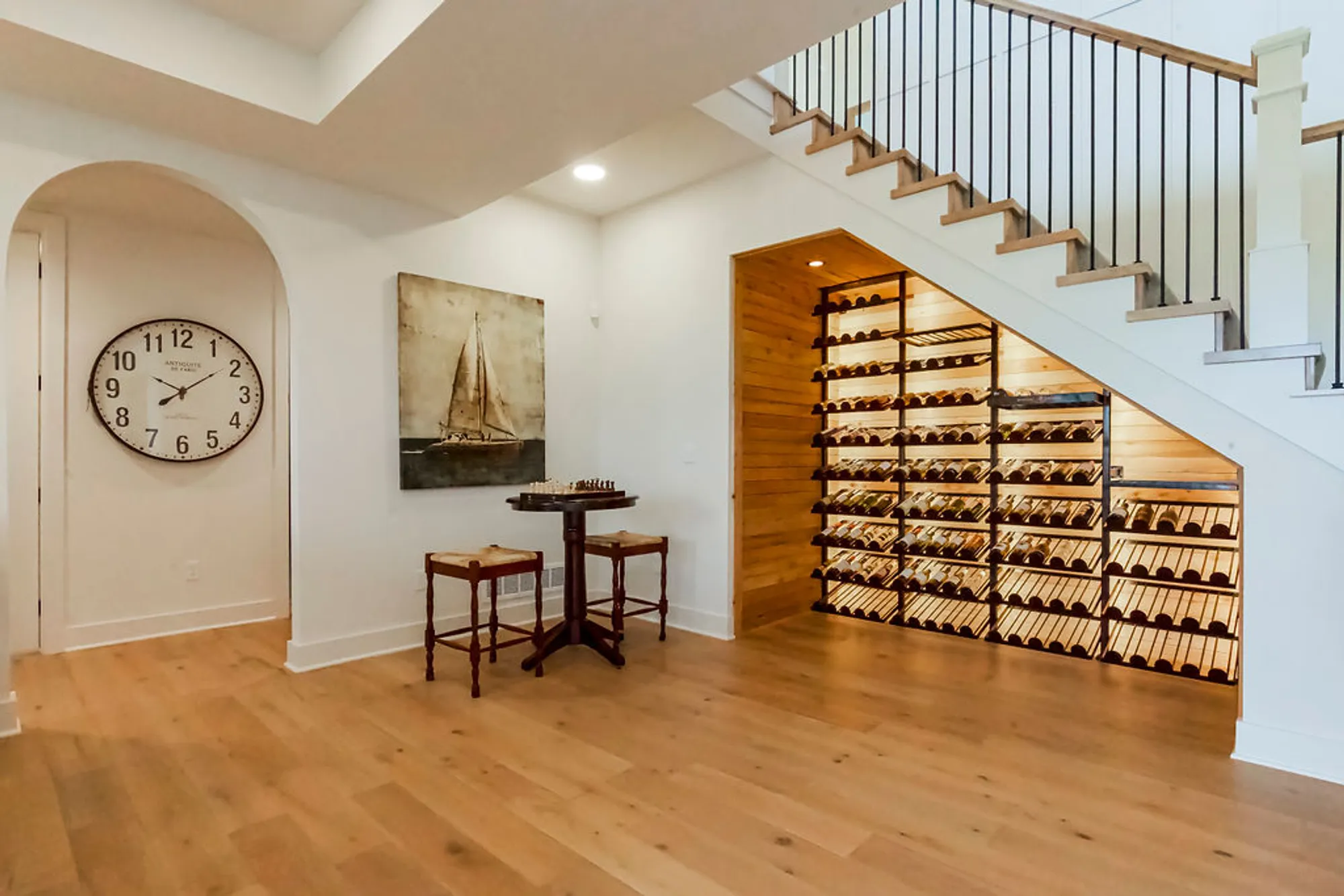 Living area with a wine rack under the staircase, a large wall clock, a sailboat painting, and a round table with two stools on wooden floors.