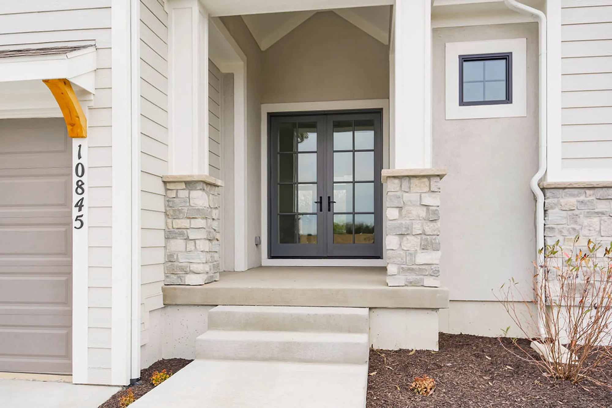 Front entry porch of a modern house featuring a gray double door with grid panes, stone pillars, and concrete steps.