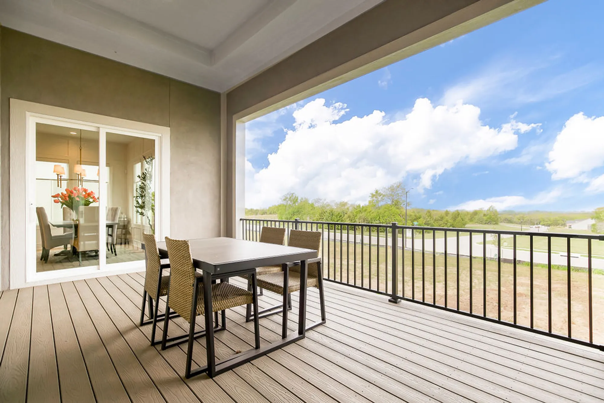 Outdoor balcony dining area with a rectangular table and wicker chairs, sliding glass doors to an interior dining room, and a blue sky with clouds.
