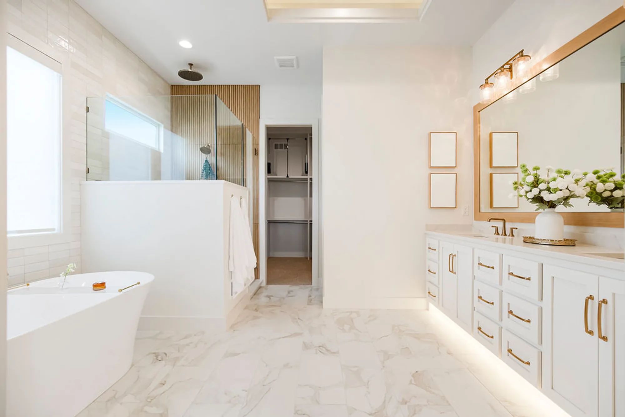 Bright modern bathroom with a freestanding tub, marble floors, glass shower, and white vanity with gold hardware; flowers on the counter.