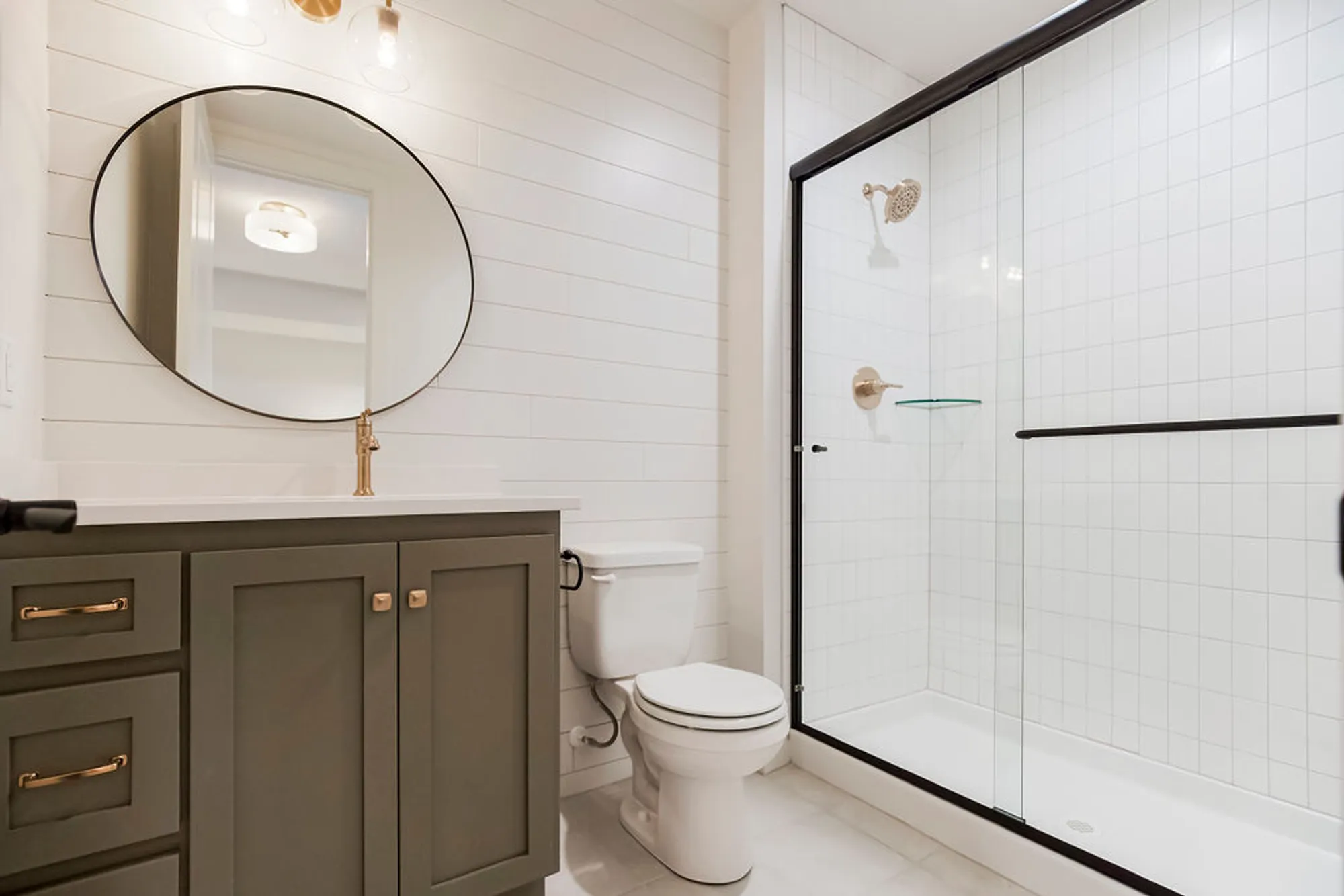 Modern bathroom with a gray vanity, round mirror, and gold faucet, white subway-tiled walls, and a glass shower with a black frame.