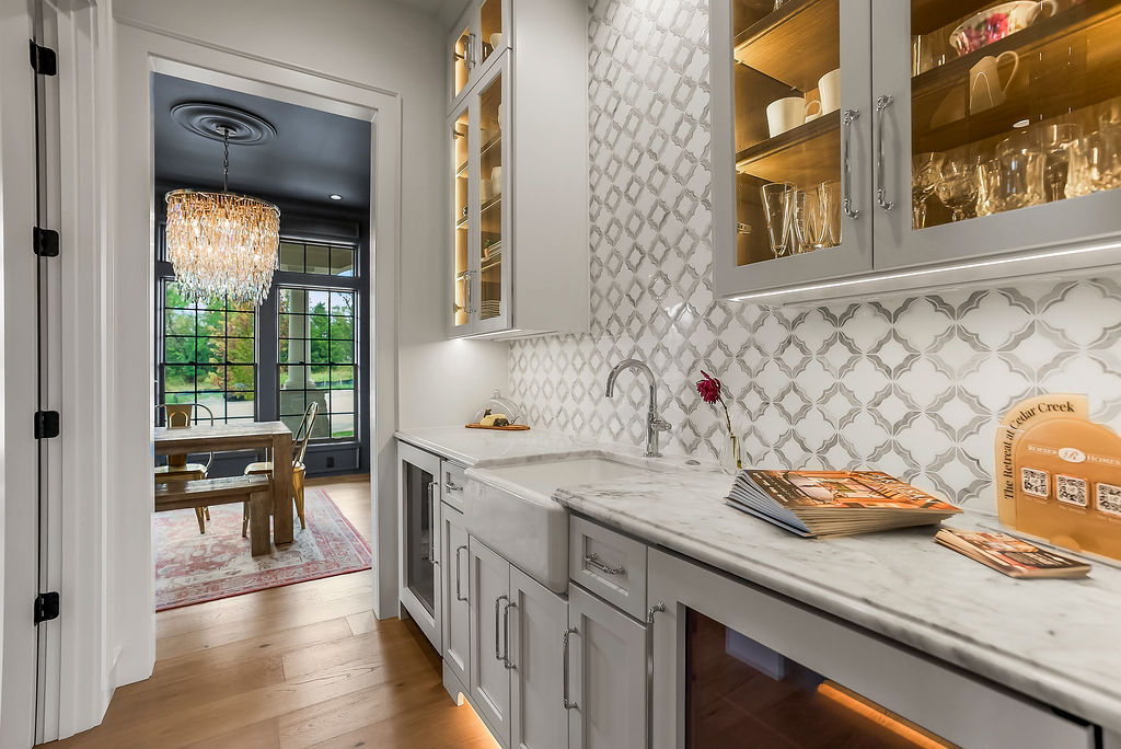 White kitchen with marble countertop, glass-front cabinets, and a patterned backsplash; doorway opens to a chandelier-lit dining room.